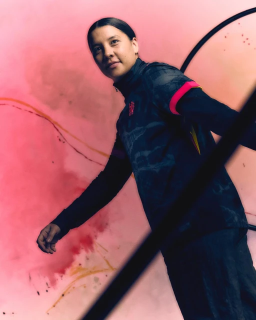 An athletic woman in a dark long-sleeved shirt poses against a pink background with splashes of dark paint-like textures and graphic lines.