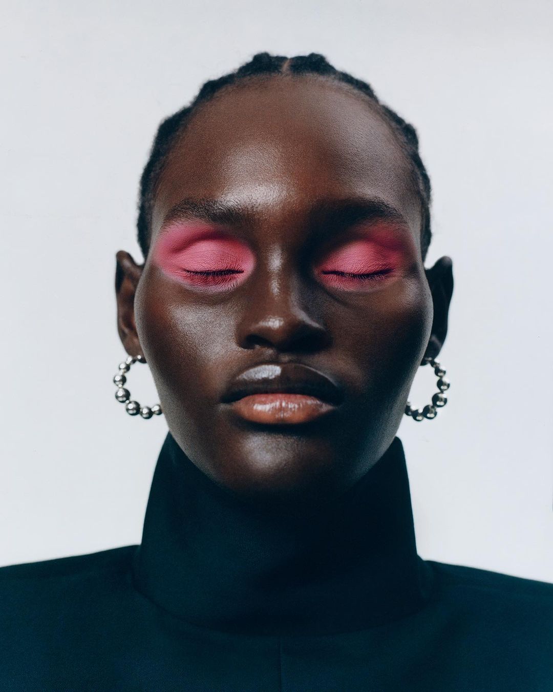 Close-up beauty portrait of a Black woman with bright pink eyeshadow, beaded silver hoop earrings, and a black high-collar garment.