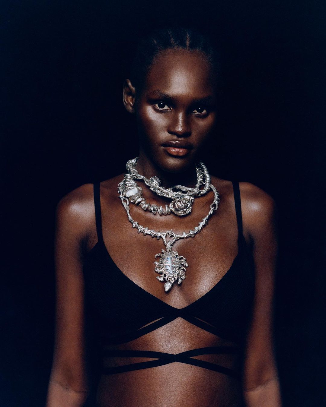 Black woman wearing a black top and layered silver statement necklaces against a dark background