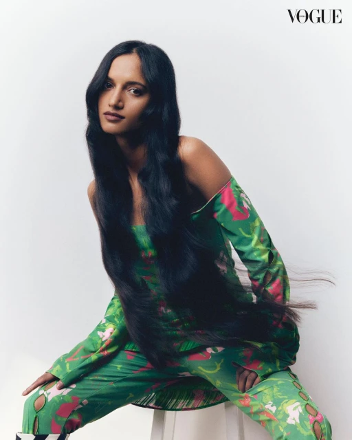 A South Asian woman poses indoors with long dark hair, wearing a green patterned two-piece outfit and high heels. Vogue logo visible.