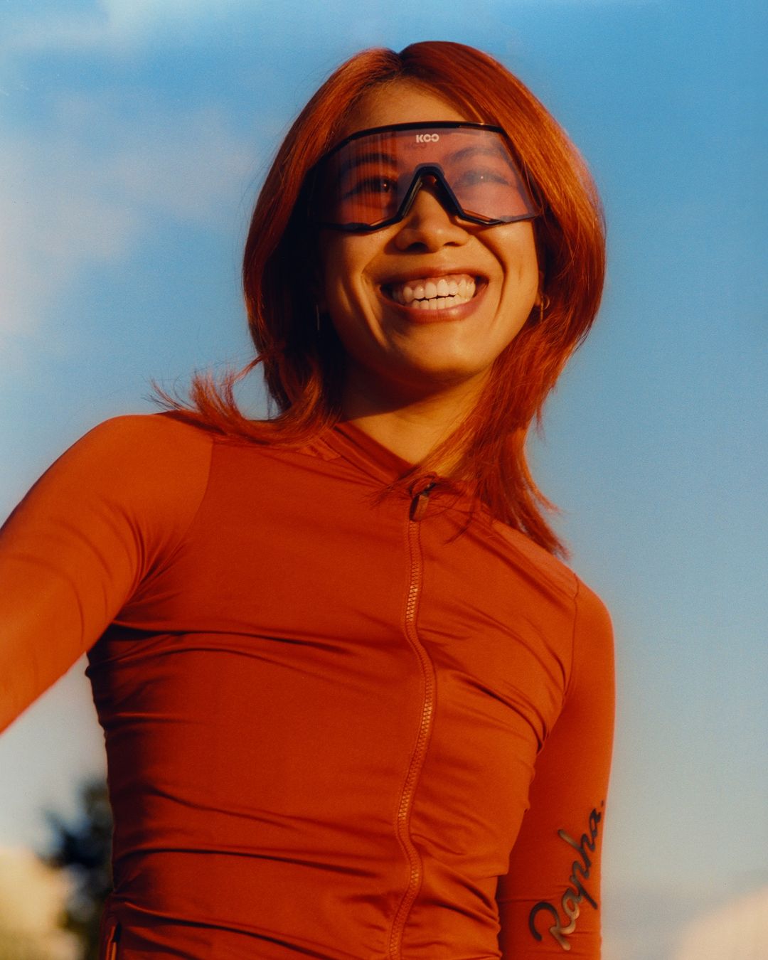 Smiling woman in cycling gear with orange hair and sunglasses against a blue sky, wearing a long-sleeved orange cycling jersey.