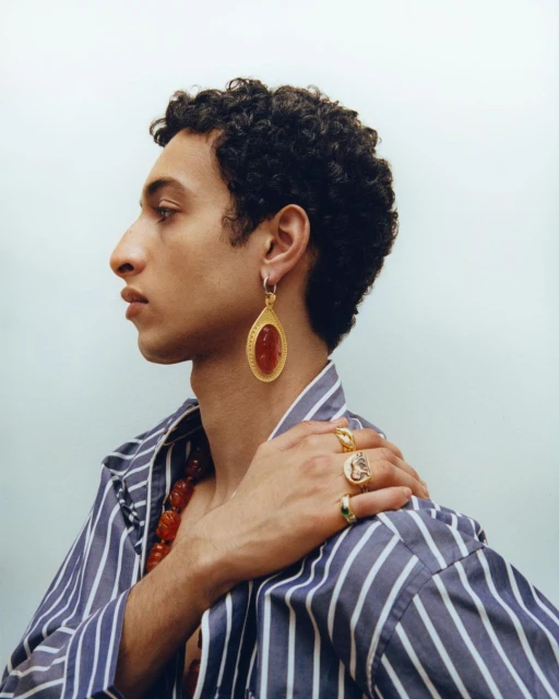 Profile of a curly-haired person in a striped shirt, accented with gold jewelry: large earrings, rings, and a red beaded necklace.
