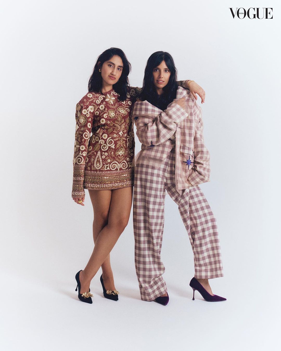 Two women pose indoors against a white background, showcasing a brown patterned mini dress and a pink plaid suit with black heels. Vogue logo visible.