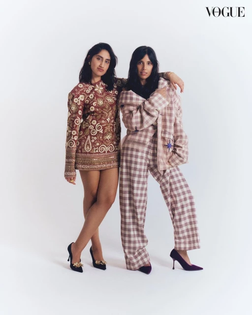 Two women pose indoors against a white background, showcasing a brown patterned mini dress and a pink plaid suit with black heels. Vogue logo visible.