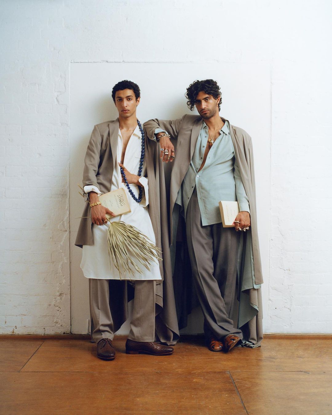 Two male models pose in tailored suits against a white wall, one holding a book and dried palms, showcasing high fashion and style.