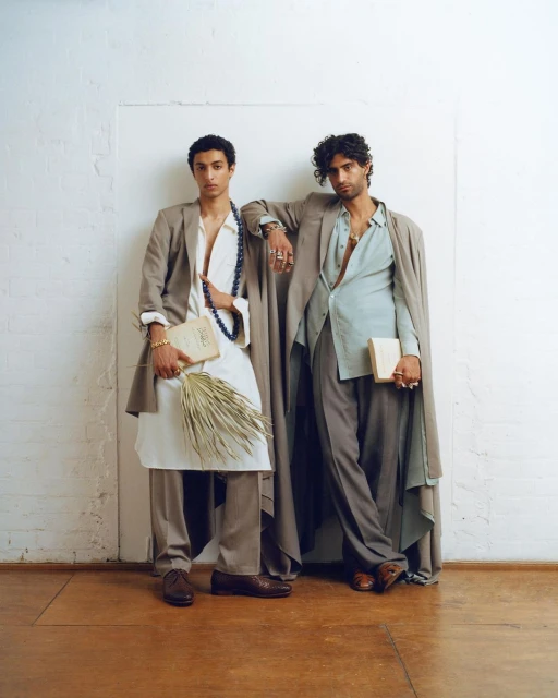 Two male models pose in tailored suits against a white wall, one holding a book and dried palms, showcasing high fashion and style.