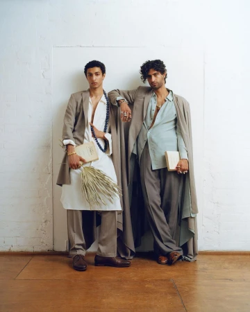 Two male models pose in tailored suits against a white wall, one holding a book and dried palms, showcasing high fashion and style.