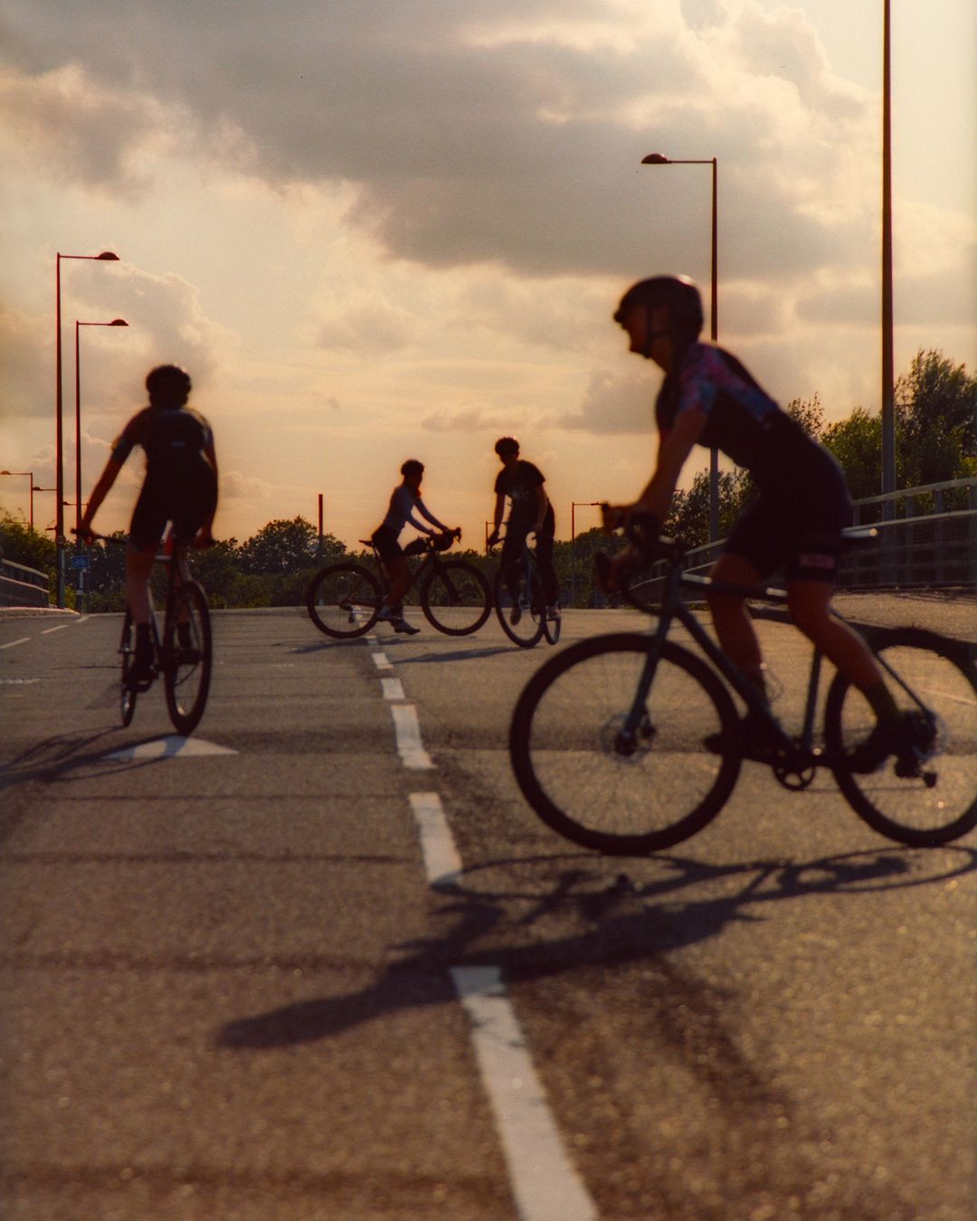 Group of cyclists riding bikes on a road with streetlights under a cloudy sky at golden hour, creating long shadows on the pavement.