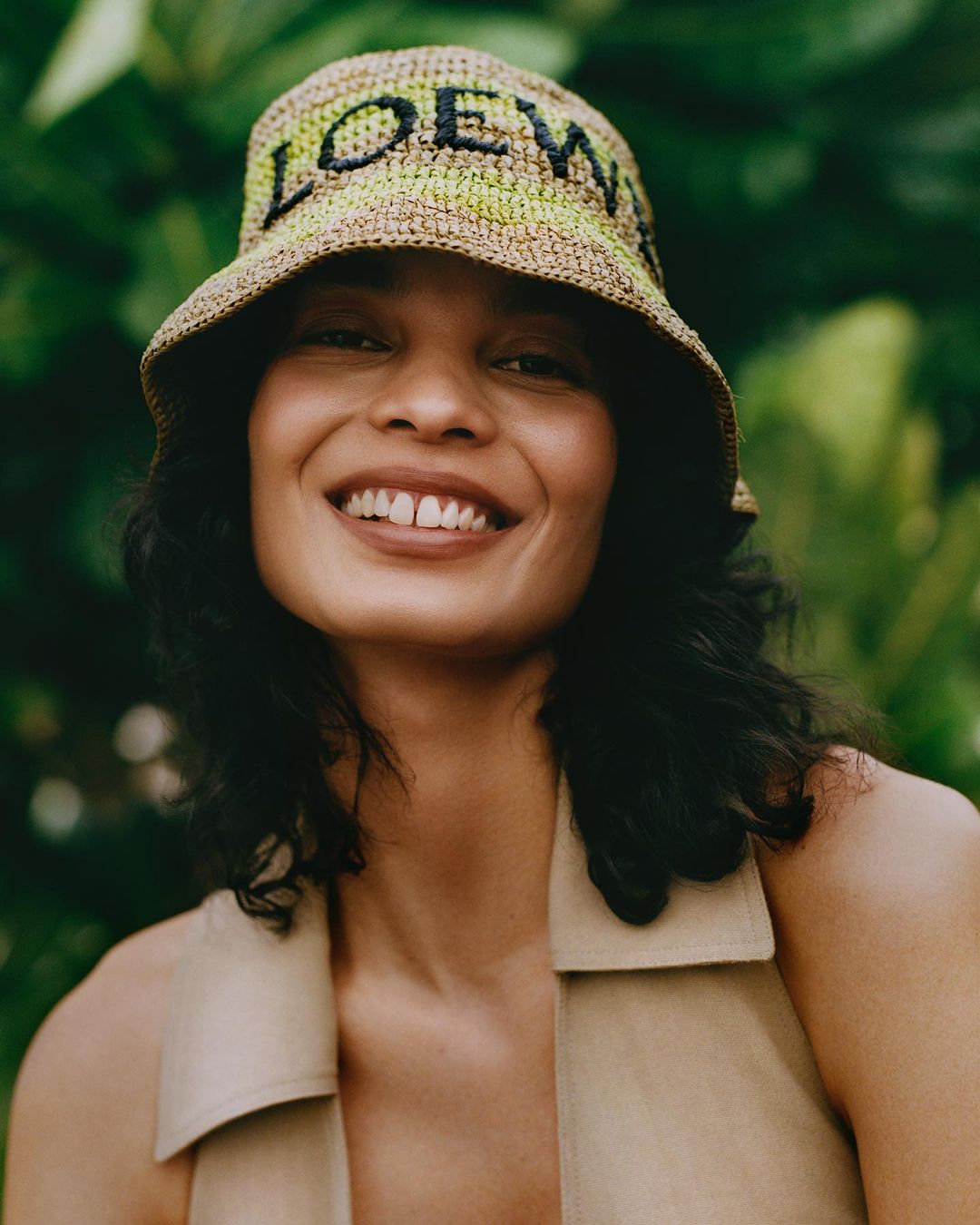 Portrait of a smiling adult woman wearing a Loewe bucket hat against a lush green leafy background in a close-up shot.