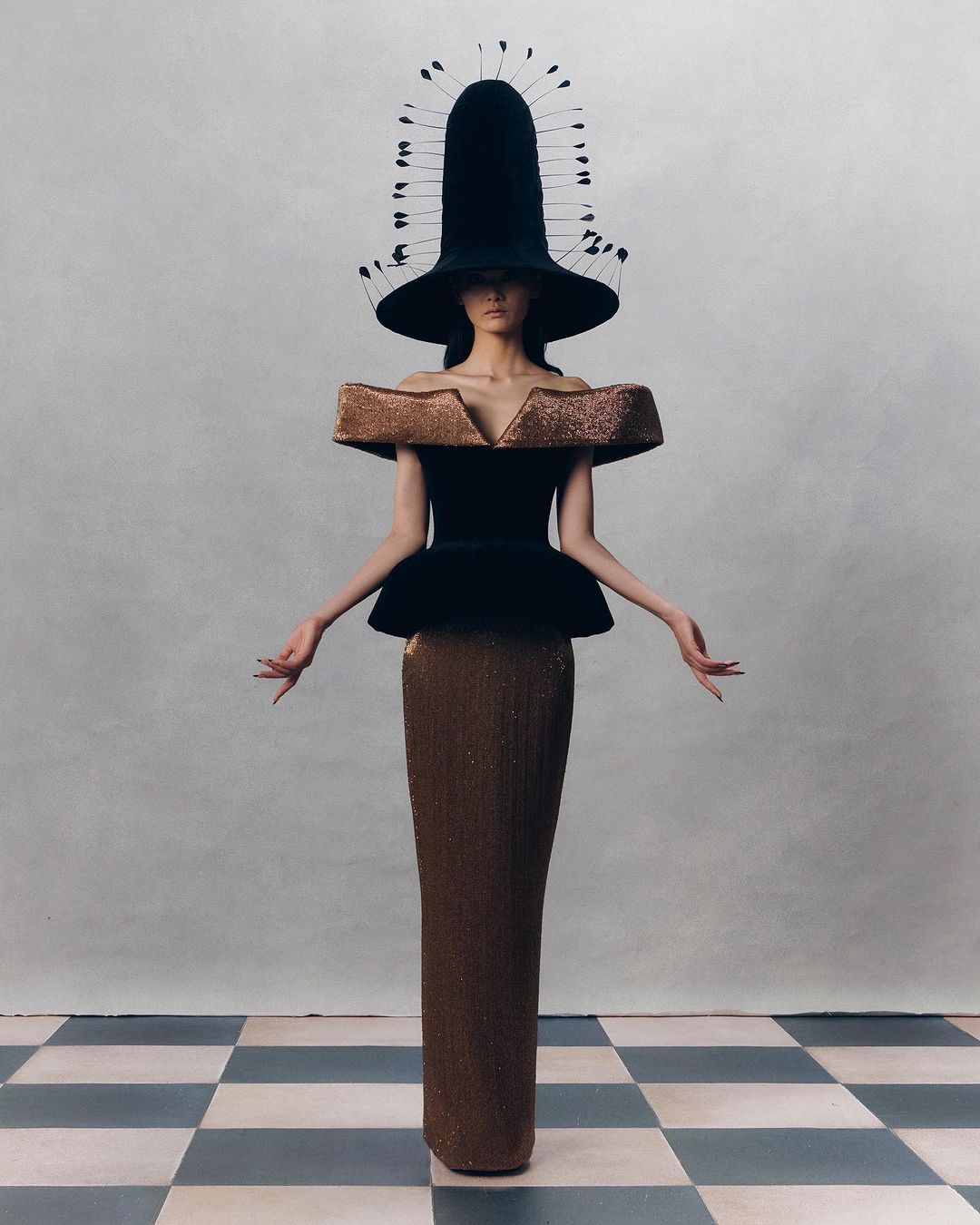 Fashion portrait of a woman wearing a large hat and gown with geometric textures, posing in front of a neutral wall with a checkered floor.
