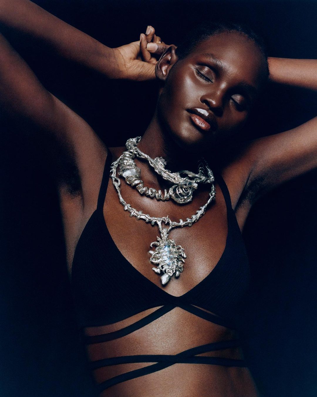 Close-up of a dark-skinned woman with a silver necklace and black strappy top against a dark background, showcasing fashion and beauty.