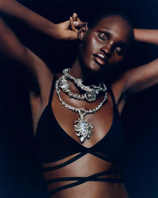 Close-up of a dark-skinned woman with a silver necklace and black strappy top against a dark background, showcasing fashion and beauty.