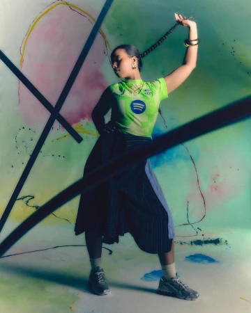 Young woman poses confidently in a fashion shot, wearing a green sports jersey, pleated skirt, and sneakers against an abstract backdrop.
