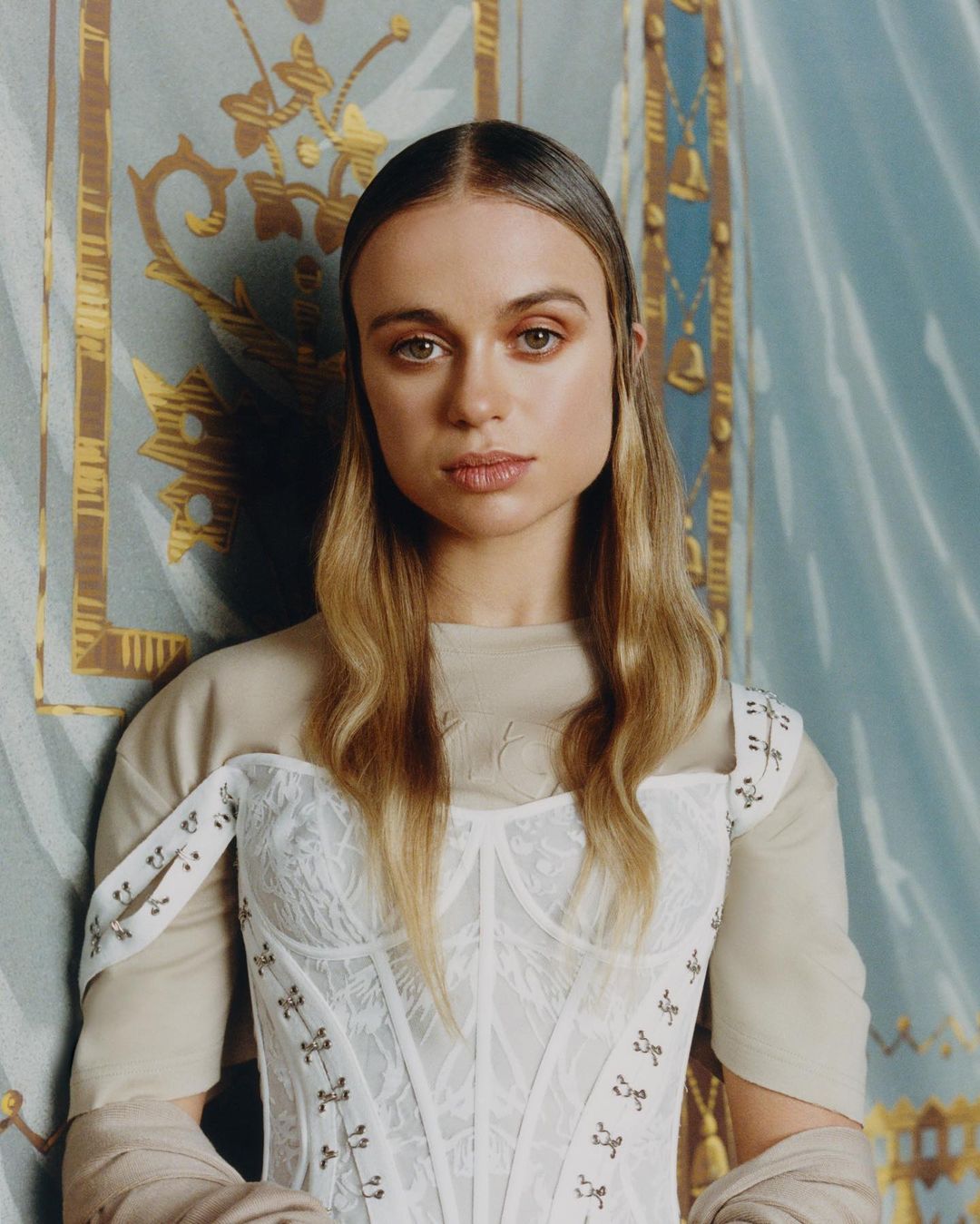 Portrait of a woman with fair skin and light brown hair, wearing a white corset top, against a patterned light blue backdrop.