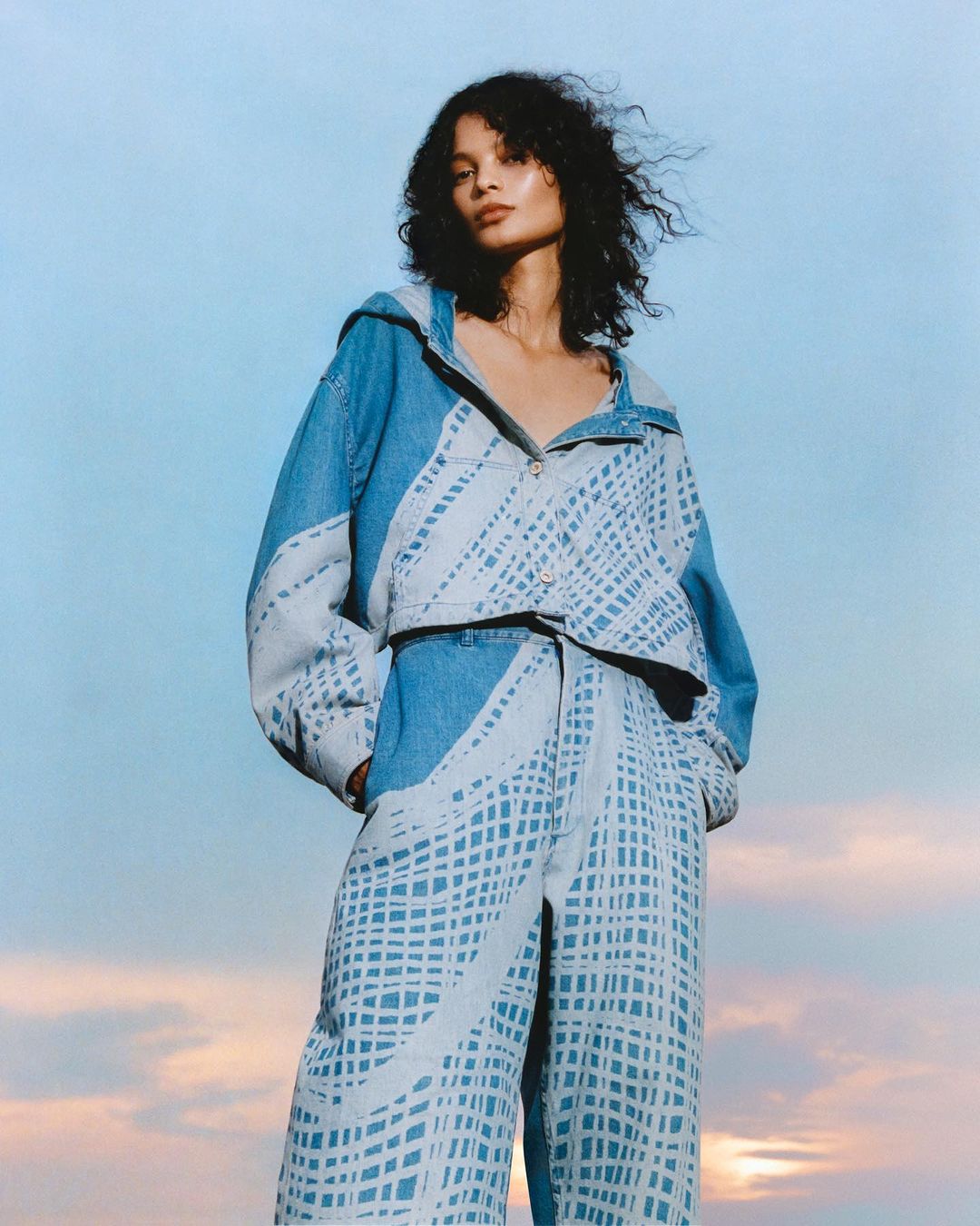 Adult female model in printed denim hooded jacket and matching pants poses against a blue sky with sunlit clouds at the horizon.