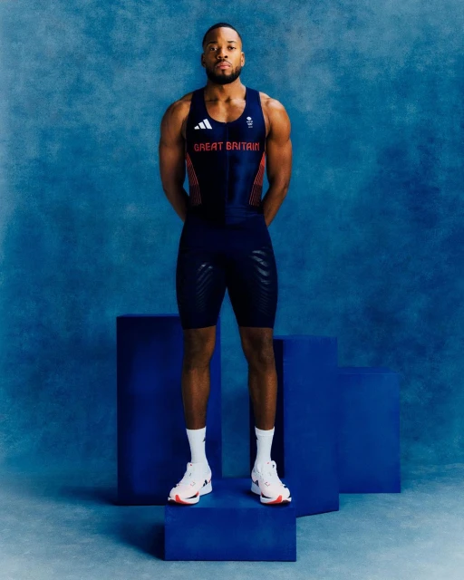 An adult male athlete stands on a royal blue platform, wearing a navy blue and red uniform, in front of a textured light blue background.