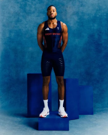 An adult male athlete stands on a royal blue platform, wearing a navy blue and red uniform, in front of a textured light blue background.