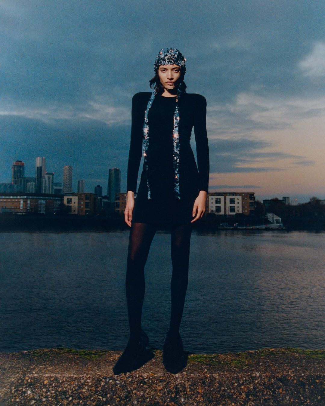 Full-length shot of a woman in a black dress, tights and shoes, wearing a patterned headscarf and scarf, with a cityscape background.