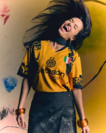 Portrait of a young adult woman in a yellow soccer jersey with long dark hair and open mouth, smiling widely in a casual setting.