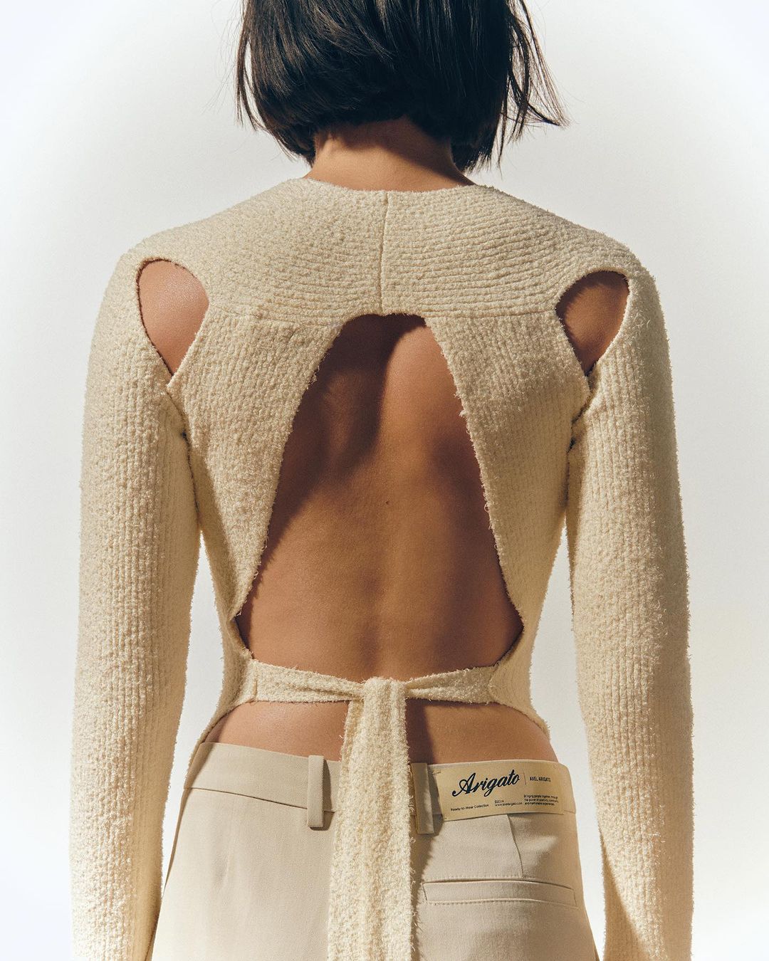 A woman with short dark hair wears a cream-colored, backless, long-sleeved sweater and khaki pants against a white background in a studio shot.
