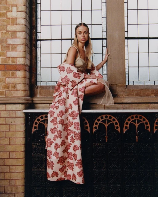 Elegant woman with long blonde hair posing in a floral coat and dress in front of a tall window with brick and dark marble walls.