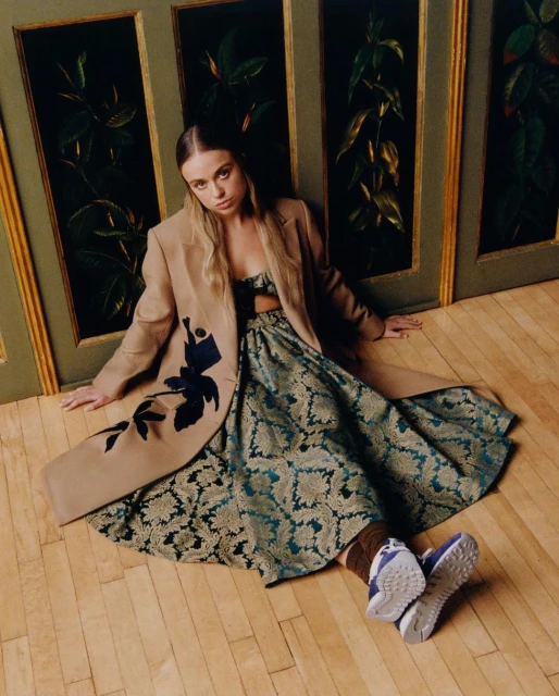 Blonde-haired woman wearing an embroidered jacket and dress sitting on a wood floor in front of green and gold wall panels.