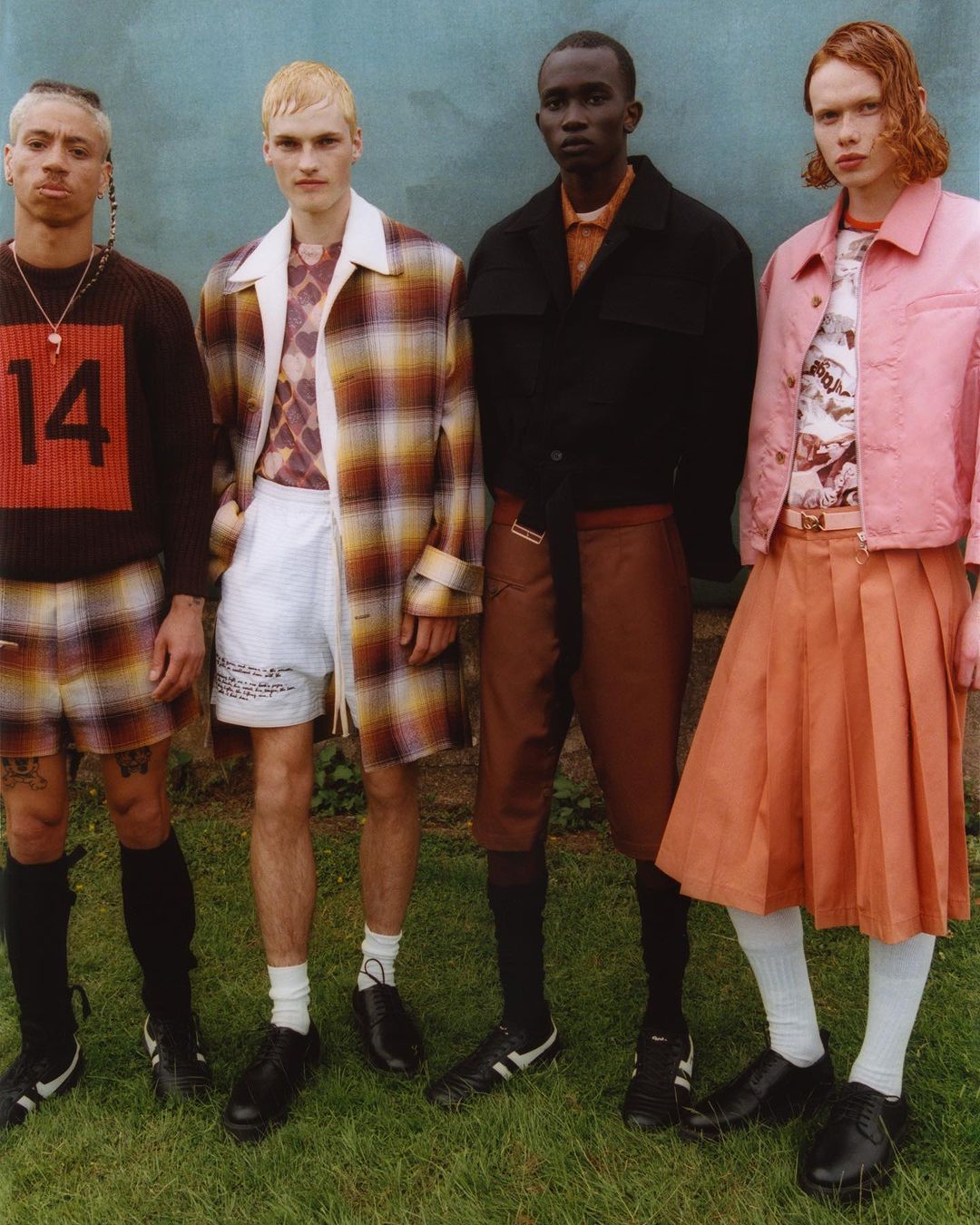 Four fashion models in varied outfits stand outdoors against a muted blue wall, showcasing a mix of modern and retro styles.