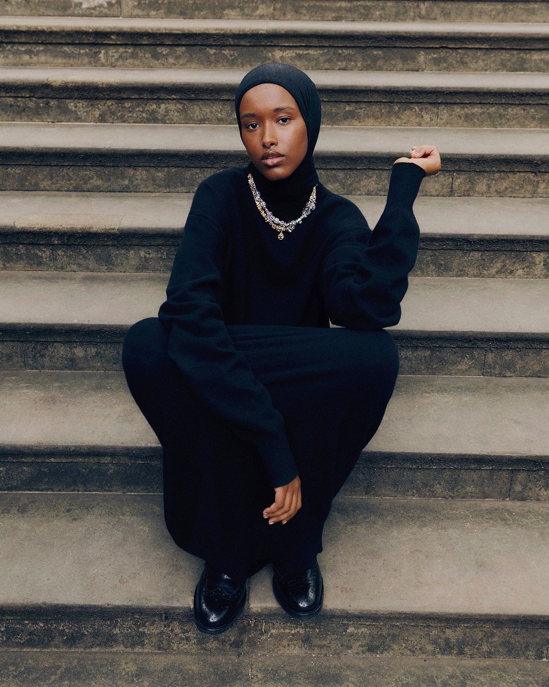Woman in black dress and hijab sits on stone steps, accessorized with a layered silver pendant necklace and dark shoes.