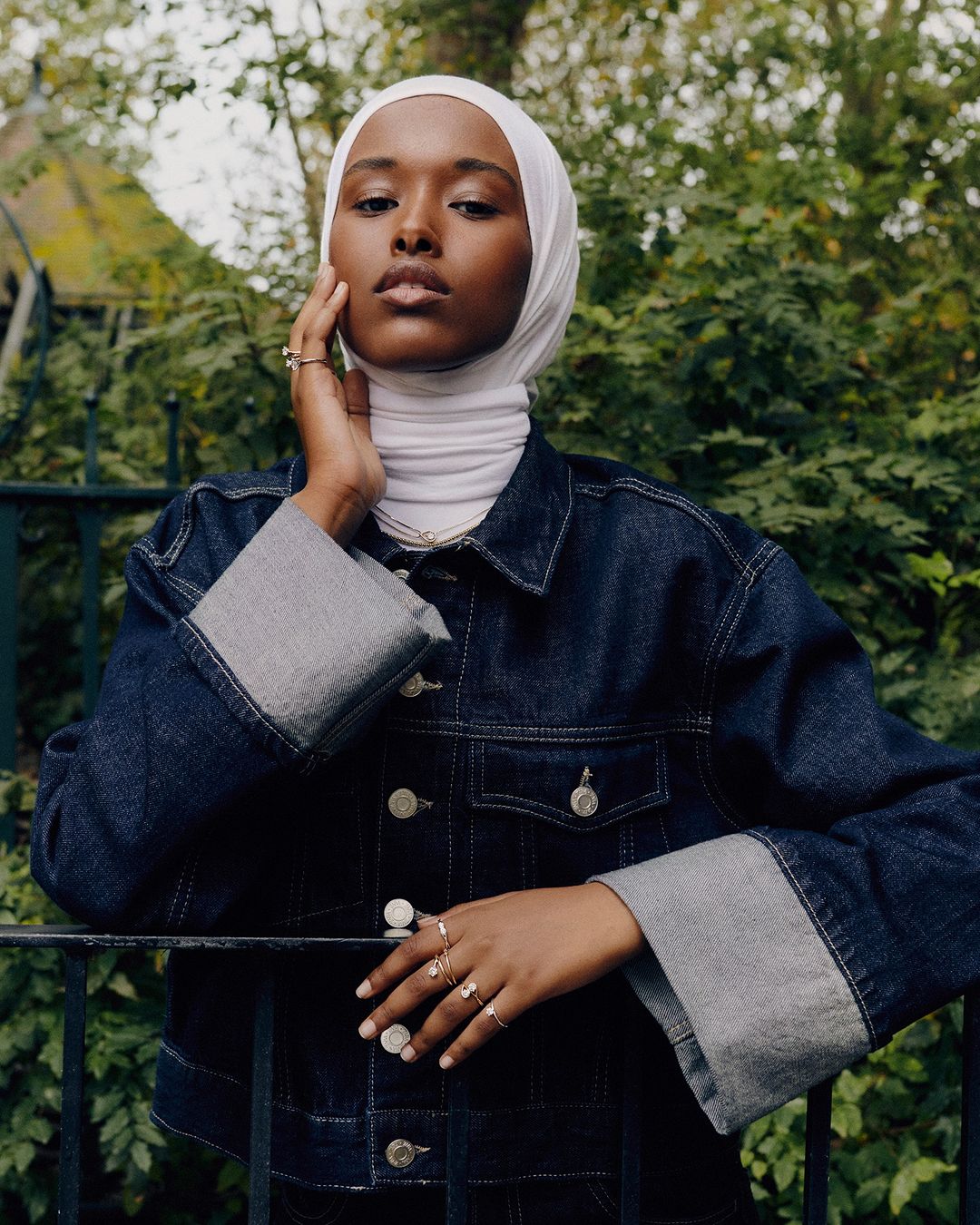Woman in denim jacket and white hijab poses outdoors, hand to face, adorned with rings, capturing a fashion-forward editorial portrait.