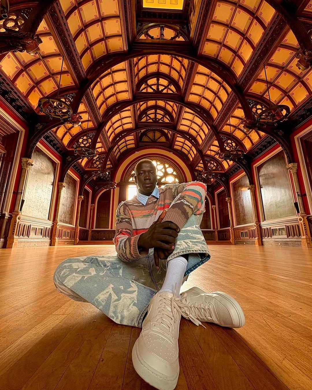 A man sits cross-legged in an ornate hall. He wears a patterned sweater and jeans, facing the camera in a wide shot.
