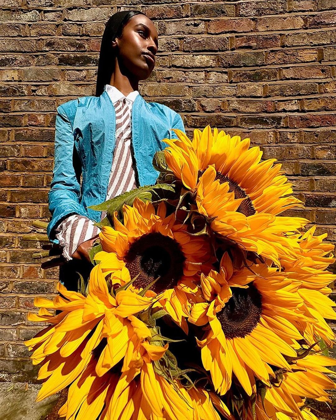 Stylish adult female posing with a bouquet of vibrant sunflowers against a textured brick wall in natural outdoor light.
