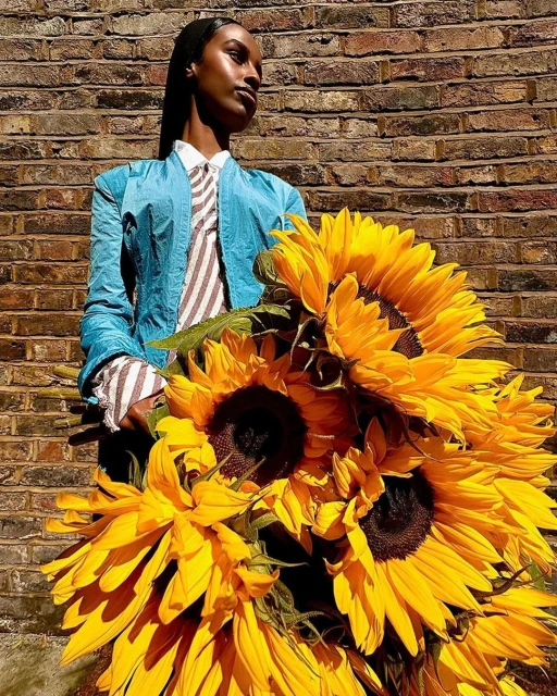 Stylish adult female posing with a bouquet of vibrant sunflowers against a textured brick wall in natural outdoor light.