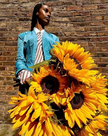 Stylish adult female posing with a bouquet of vibrant sunflowers against a textured brick wall in natural outdoor light.