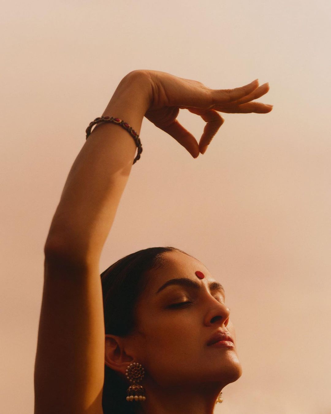 Woman in profile with hand raised, wearing a bracelet and earrings against a peach-colored sky. Eyes closed, red dot on forehead.