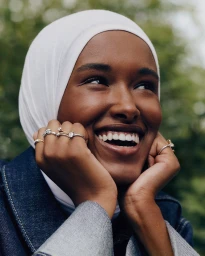 Close-up portrait of a smiling Black woman wearing a white hijab and denim jacket, showcasing multiple rings on her fingers.
