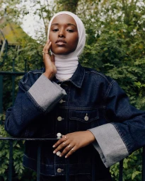 Portrait of a Black woman wearing a white hijab and dark denim jacket leaning against a black metal fence outdoors.