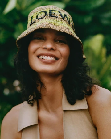 Close-up portrait of a smiling woman wearing a woven bucket hat with black lettering and a beige sleeveless top.
