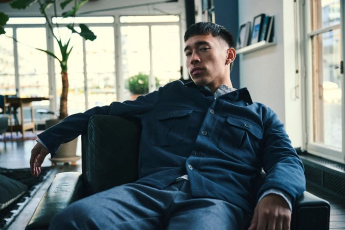 Young man in a blue chore coat and gray pants sitting indoors on a black leather couch in front of bright windows.