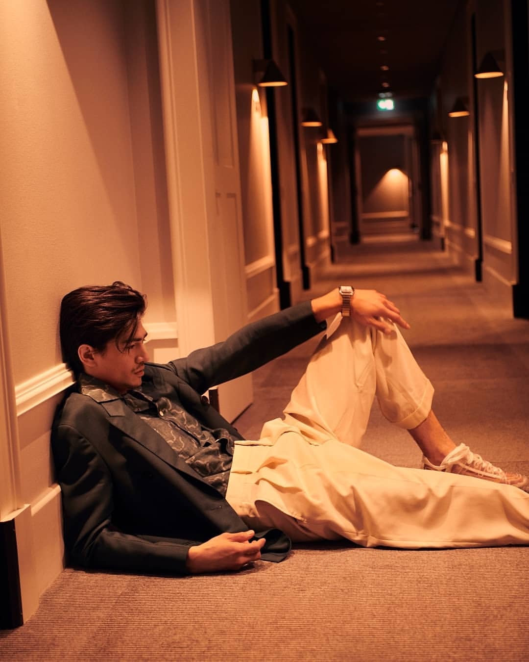 Stylish adult male in blazer and beige pants poses on carpeted hallway floor with lit sconces and dark end. Indoor fashion shot.