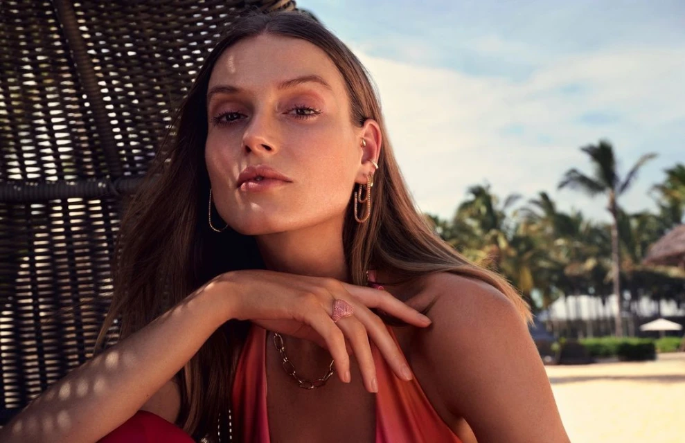 Adult female with jewelry posing in front of palm trees and sky. She has brown hair and is wearing a peach-colored top and gold jewelry.