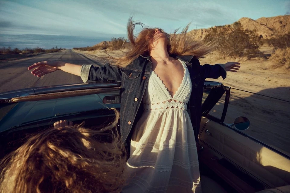 Two women in a convertible car on a desert road, one standing with outstretched arms, wearing a white dress and jean jacket.