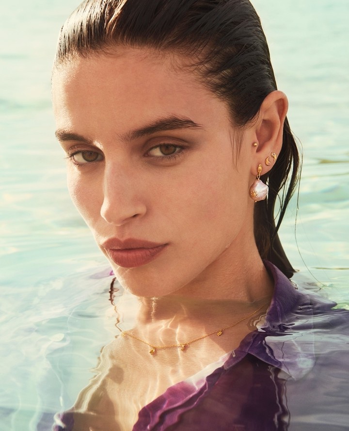 Close-up of a brunette adult woman in water wearing a purple shirt and pearl jewelry, including earrings and a delicate necklace.
