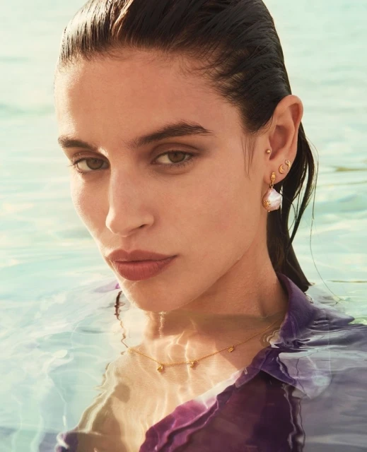 Close-up of a brunette adult woman in water wearing a purple shirt and pearl jewelry, including earrings and a delicate necklace.