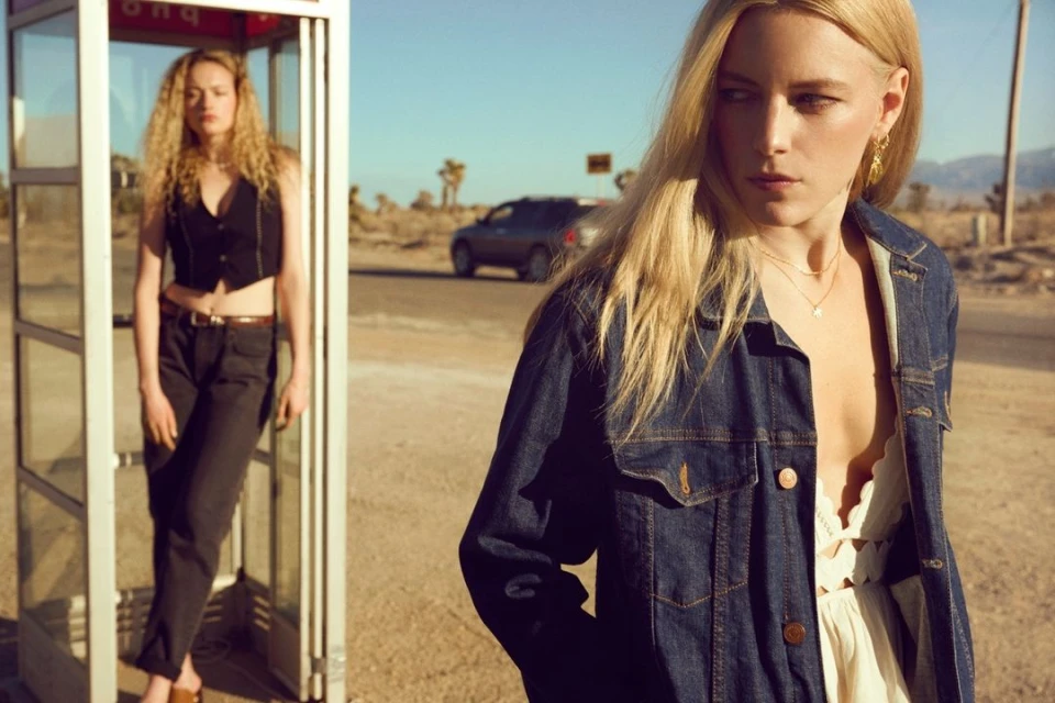 Two young women in a desert setting, one posing in a phone booth, the other standing with a denim jacket, on a sunny day.