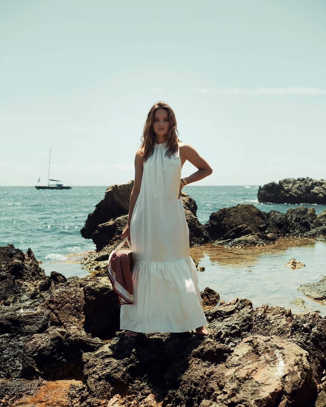 Adult female stands on rocky shore wearing a white dress, holding a bag, ocean with sailboat visible against a blue sky backdrop.