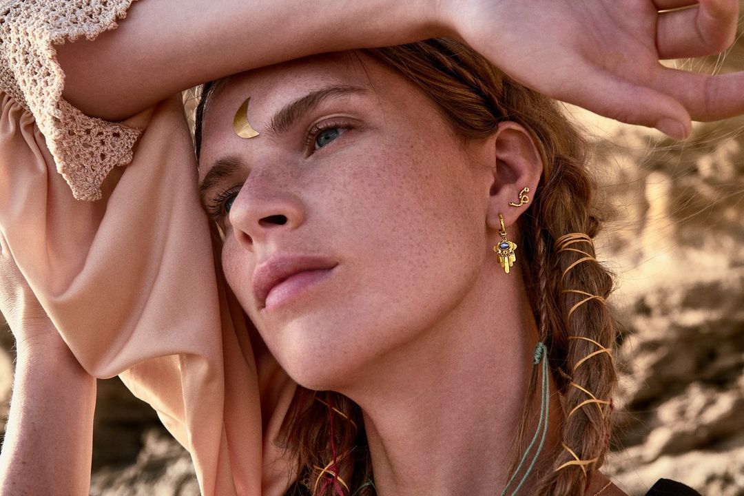 Portrait of a fair-skinned woman with freckles wearing gold jewelry, including a moon on her forehead, looking off to the side.