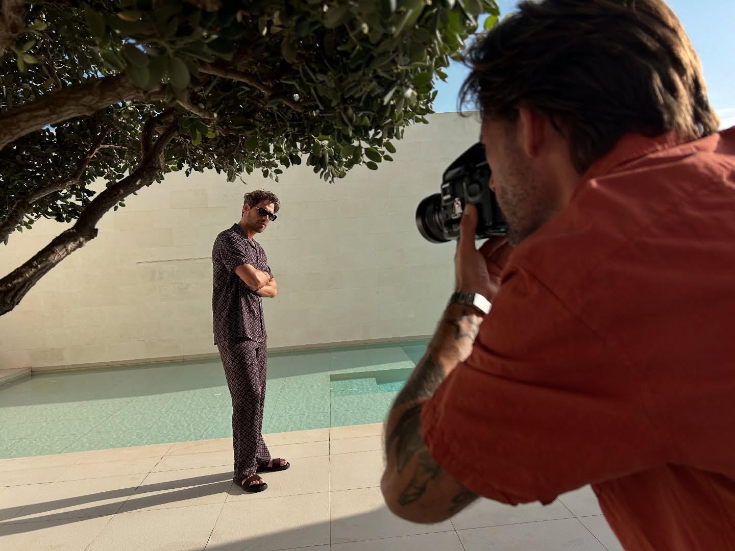 A man poses in pajamas by a pool while a photographer takes his picture outdoors during the day under a tree.