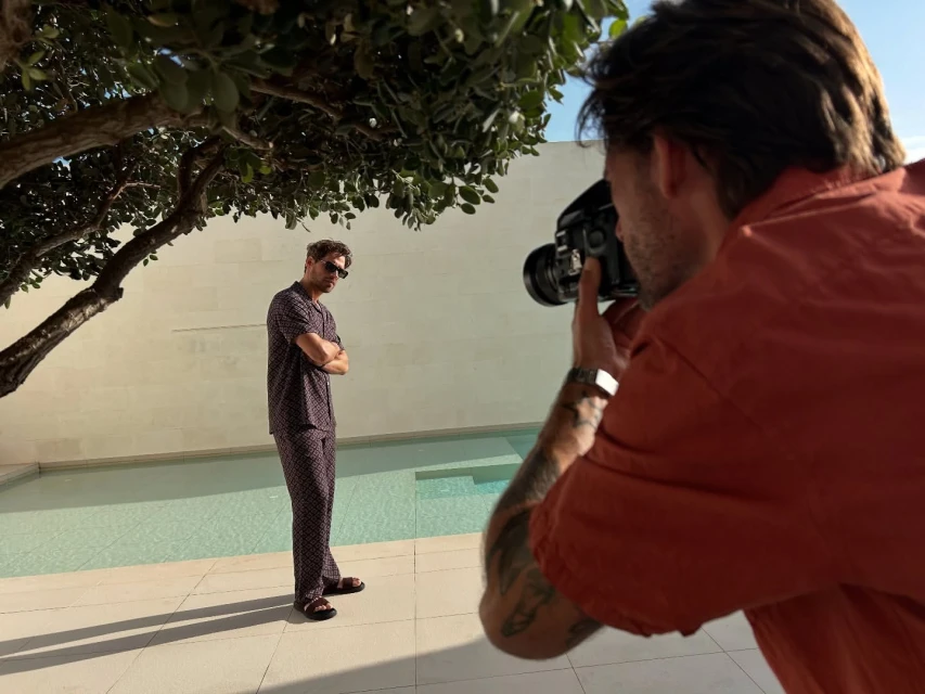 A man poses in pajamas by a pool while a photographer takes his picture outdoors during the day under a tree.