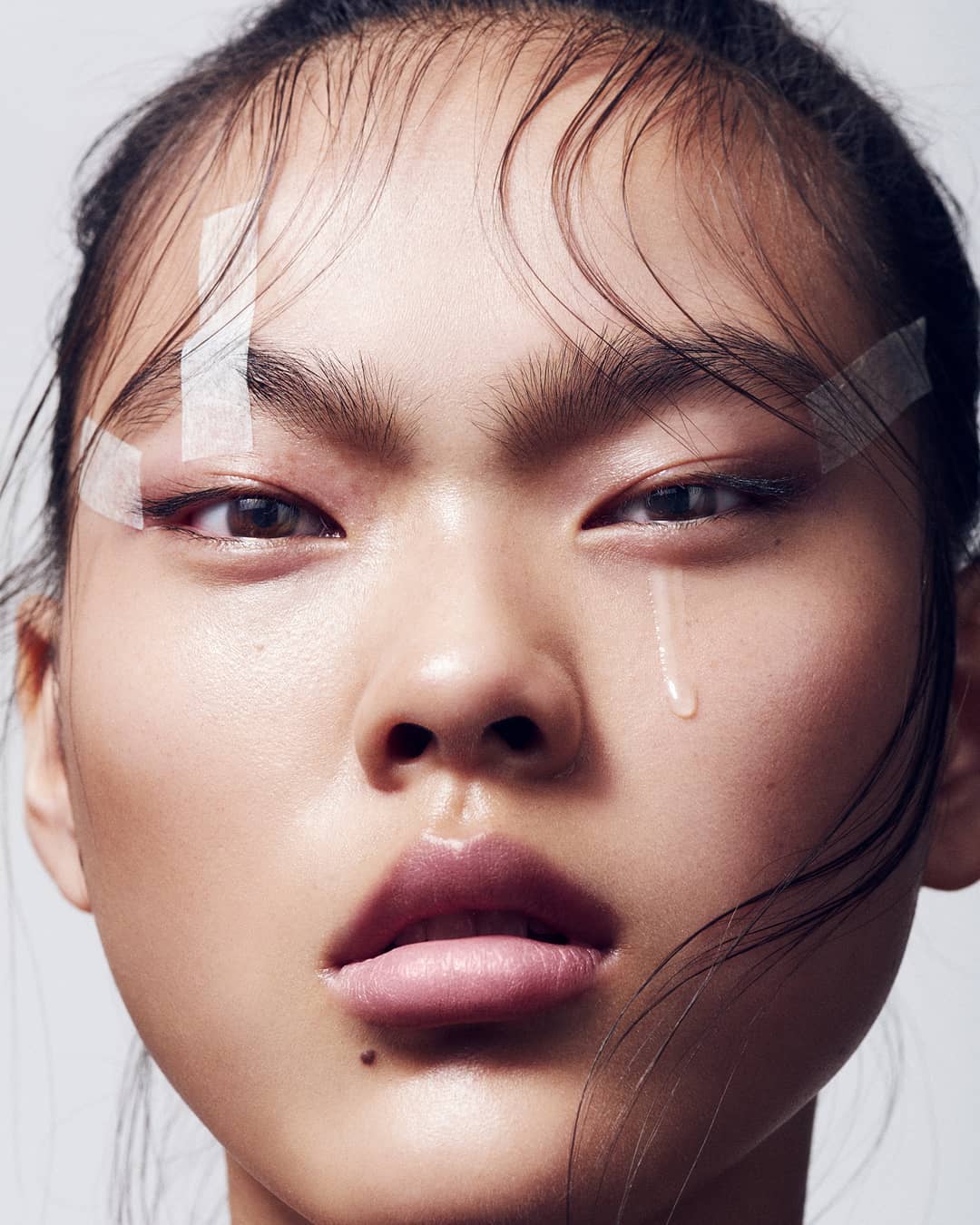 Close-up portrait of an Asian woman with a teardrop and clear tape on her forehead, highlighting beauty and makeup in a minimal setting.
