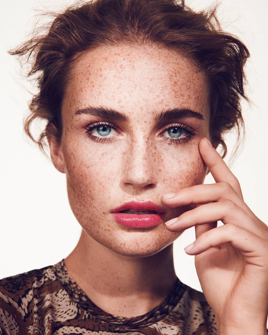Close-up portrait of a freckled woman with blue eyes and pink lipstick, posing in front of a white backdrop in a beauty fashion shoot.